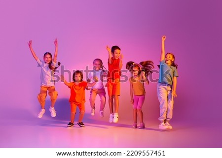 Modern choreography. Dance group of happy, active little girls and boys in bright clothes in action isolated on pink background in neon. Concept of emotions, music, fashion, art, childhood, hobby.