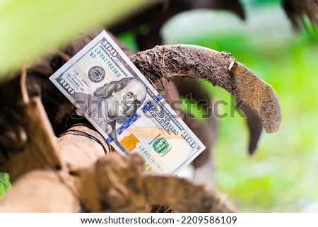 Hundred dollar bill between the teeth of a tractor rototiller close-up. Finance and agricultural machinery. Money on the immobilized tractor units. Stopping the work of the farm