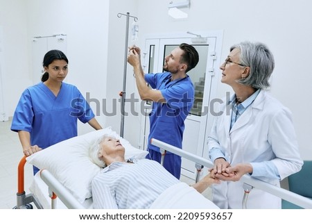 Nurse putting drip on elderly patient lying on gurney while being examined by experienced woman doctor. Medical assistants will take injured woman to her medical ward Royalty-Free Stock Photo #2209593657
