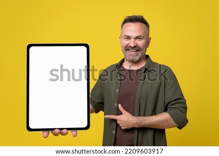Handsome bearded mature man holding huge digital tablet with white mock-up screen and pointing by hand, Man dressed in green shirt isolated on yellow background. Free space mock up.