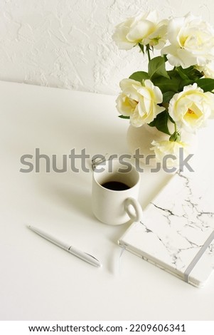 Feminine lifestyle composition, minimal workspace neutral colors. Vase with beige roses flowers, cup coffee,diary,pen on white desk near  white background with copy space .Breakfast stationary mockup.