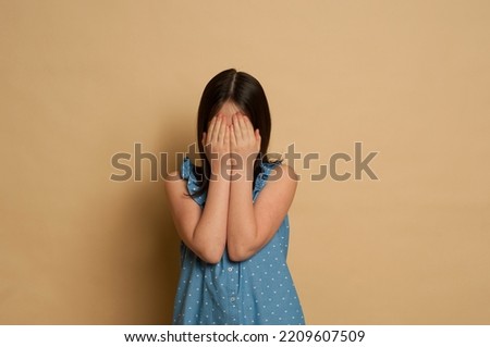 dark-haired girl 9-12 years old in a blue dress on a beige background covers her face with her hands, chagrin