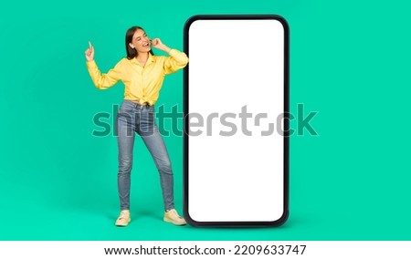 Smiling european young woman in headphones have fun, sing, enjoy music near big smartphone with blank screen, isolated on green background, studio. Ad and offer, website and new app for relaxation