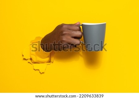 Black male hand holding a mug of coffee through torn yellow paper background, closeup, copy space. Have a break and refreshing drink Royalty-Free Stock Photo #2209633839