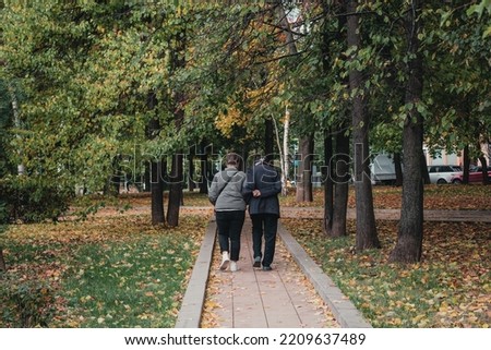 A couple of pensioners walk along a paved path in an autumn park. View from the back. Yellowing and falling foliage on trees. Yellow dry leaves on green grass. Moscow, Russia