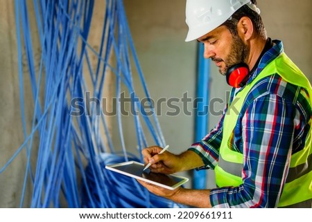 An experienced electrical engineer who checks the cables in the building. He uses a tablet to help with work. Royalty-Free Stock Photo #2209661911