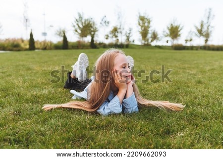 A happy little girl, in a denim jacket, lies on the green grass, lawn. a smiling child is resting, lying on the grass. the concept of a carefree childhood.