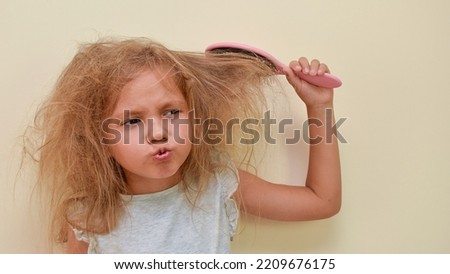 A child with very tangled hair is trying to comb his hair. A little girl's hair is hard to comb, disheveled and with tangles after washing. Hair does not comb without a conditioner balm. Royalty-Free Stock Photo #2209676175