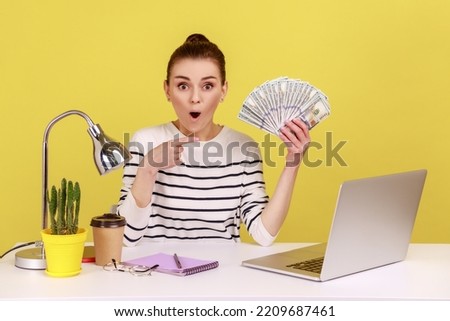 Amazed shocked woman pointing finger at hundred dollar bills, holding sitting at workplace with laptop, high salary, bonuses and perks. Indoor studio studio shot isolated on yellow background. Royalty-Free Stock Photo #2209687461