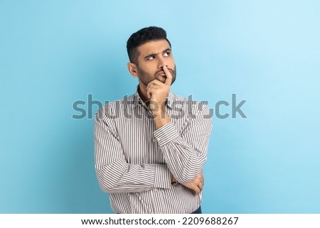 Portrait of thoughtful handsome bearded businessman holding his chin and pondering idea, confused not sure about solution, wearing striped shirt. Indoor studio shot isolated on blue background. Royalty-Free Stock Photo #2209688267