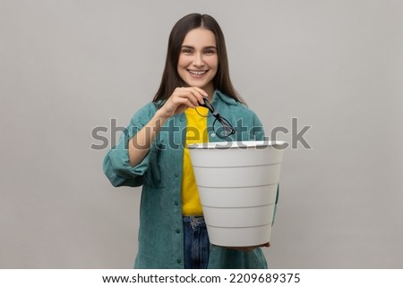 Smiling woman with dark hair standing holding trash bin and throwing out eyeglasses, improving eyesight after treatment, wearing casual style jacket. Indoor studio shot isolated on gray background. Royalty-Free Stock Photo #2209689375