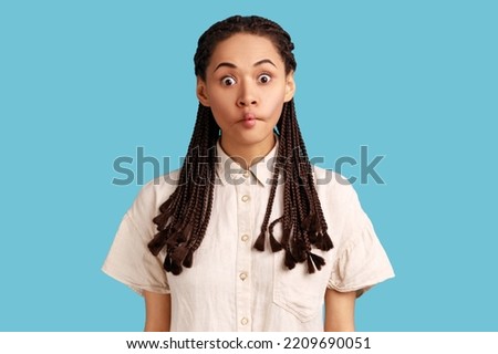 Surprised young woman with dreadlocks makes fish lips, being not scared to show emotions, tries to make her crying friend laugh, wearing white shirt. Indoor studio shot isolated on blue background.