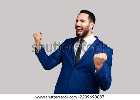 Bearded man showing yes gesture and screaming celebrating his victory, success, dreams comes true, euphoria, wearing official style suit. Indoor studio shot isolated on gray background. Royalty-Free Stock Photo #2209690087