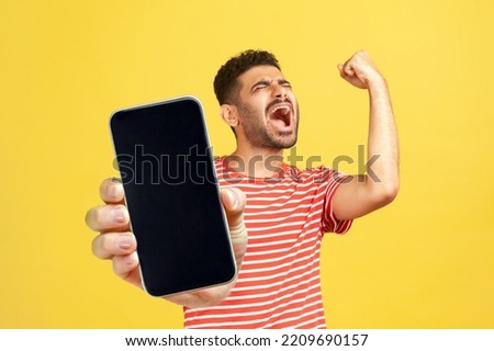 Extremely happy joyful man with beard wearing red T-shirt standing showing big mobile phone with empty display, celebrating his success, winning. Indoor studio shot isolated on yellow background. Royalty-Free Stock Photo #2209690157