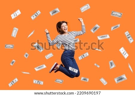 Portrait of satisfied excited woman jumping with clenched fist, celebrating winning lottery, flying in money rain, dollars falling. Indoor studio shot isolated on orange background.