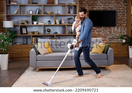 A young father, with a child, does housework. Father on maternity leave. Dad cheerfully sings, dances and vacuums, cleans with a small child in his arms. Paternity Leave Royalty-Free Stock Photo #2209690721