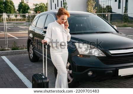 Stylish middle aged woman holding travel bag talking on mobile phone standing near new car. Business trip  concept	
 Royalty-Free Stock Photo #2209701483