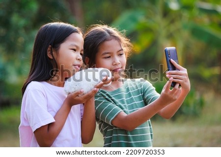 Two asian girls taking a picture and playing with adorable bunny. Sister siblings holding a little rabbit Holland lop on hand and using smartphone selfie together with fun in the garden.