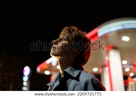 Pacified Caucasian woman in glasses is standing next to car filling station and enjoying falling snowflakes. Pretty girl with short haircut looks up into sky watching coming winter or night cold snap Royalty-Free Stock Photo #2209721775