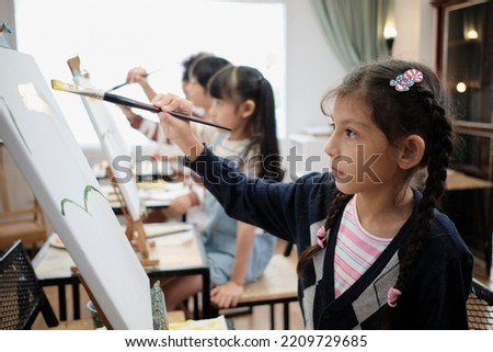 A little girl concentrates on acrylic color picture painting on canvas with multiracial kids in an art classroom, creative learning with talents and skills in the elementary school studio education.
