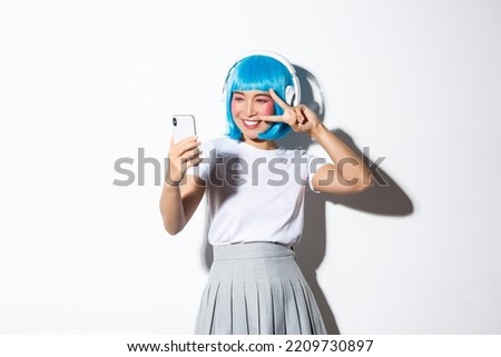 Portrait of cute asian girl dressed up as anime character, posing for selfie in headphones and blue short wig, taking picture on smartphone, standing over white background