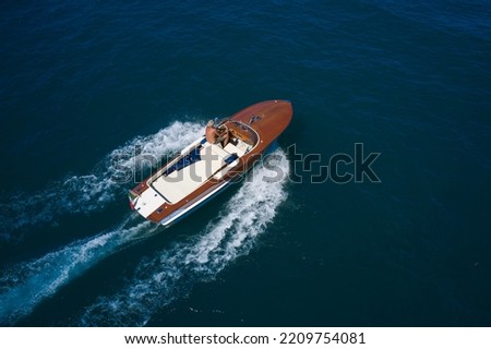 Big expensive wooden open boat with a man moving fast on dark blue water top view. Italian wooden speedboat moving up aerial view. Wooden expensive boat fast movement on dark water top view. Royalty-Free Stock Photo #2209754081