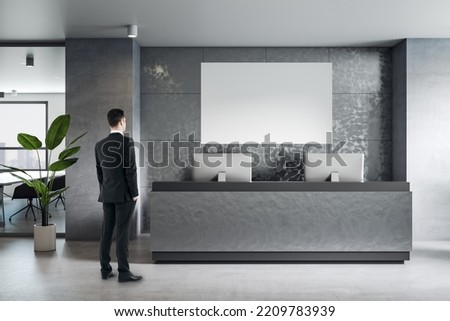 Businessman back view looking at blank white poster with space for your logo or text on grey wall in reception area with modern computers, concrete floor and green plant, mockup