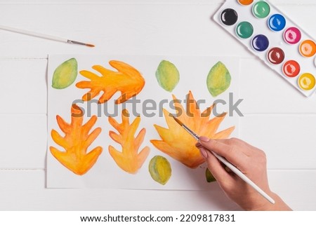 Female hand drawing autumn leaves of different shapes, sizes and colors with watercolors and brush on white wooden table. Top view. DIY handmade autumn decorations