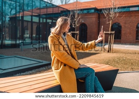 Portrait of happy woman in a yellow coat taking selfies and video calls with friends in a modern city park, smiling to camera. 