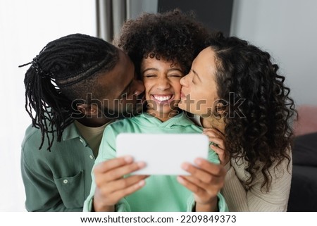 happy african american parents kissing cheek of daughter while taking selfie at home