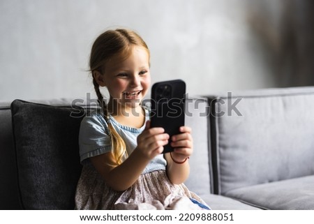 Portrait of cute little cute girl holding smartphone and resting at sofa at home. Choosing favorite music or cartoons,texting messages,browsing internet,watching video, playing games on mobile phone