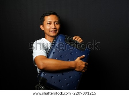 Happy Asian man is carrying a blue suitcase isolated on black background.