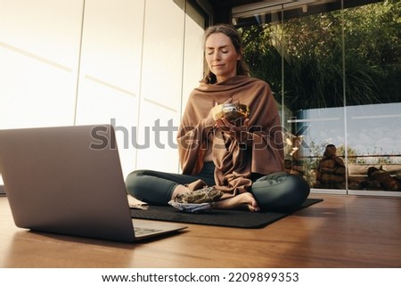 Ayurvedic healer meditating with a singing bowl and sage during an online holistic class. Woman performing a self-healing and purifying ceremony at home. Senior woman taking care of her ageing body. Royalty-Free Stock Photo #2209899353