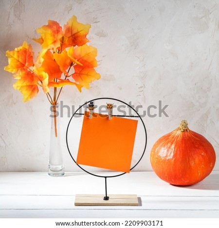 Yellow empty sheet on metal stand, orange pumpkin, decorative leaves on white. Hallowen bright decorations idea. Holiday greeting card or invitation. Copy space. Mock up. Square photo.
