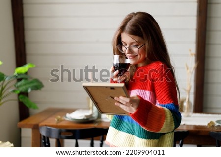 A beautiful young girl remembers a pleasant moment in her life and drinks wine in a glass. Holds a framed photograph in his hand.