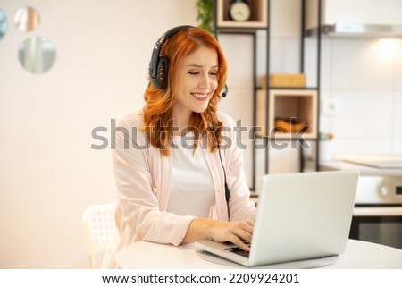 Young smiling friendly ginger girl working as agent at call center from home. Wearing headset and typing on laptop. Royalty-Free Stock Photo #2209924201