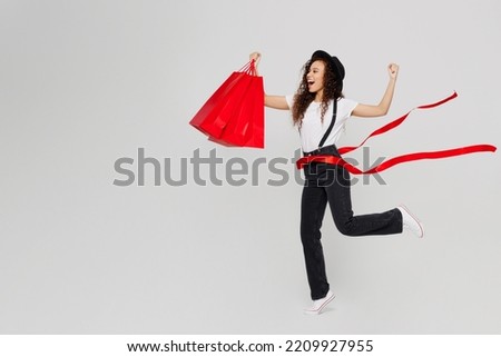 Full body exultant young woman wear t-shirt hat hold in hand paper package bags after shopping run cross red finish ribbon isolated on plain solid white background. Black Friday sale buy day concept