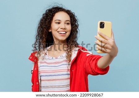 Young woman of African American ethnicity wear red jacket do selfie shot on mobile cell phone post photo on social network isolated on plain light blue cyan background. Wet fall weather season concept
