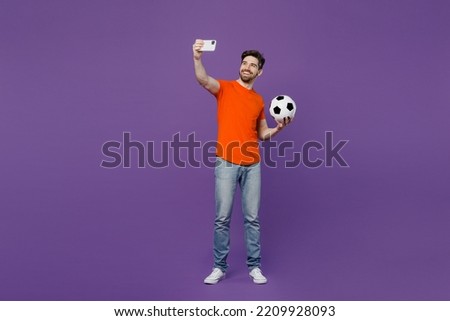 Full body young fan man he wear orange t-shirt cheer up support football sport team hold soccer ball watch tv live stream do selfie shot on mobile cell phone isolated on plain dark purple background