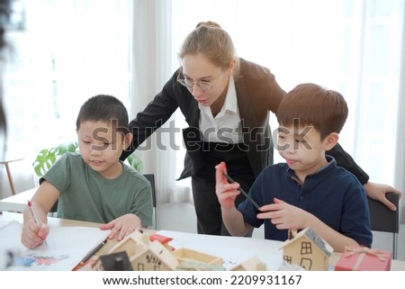 Asian lovely little boy enjoy drawing a picture and painting on the paper in art classroom. Teacher looking at the boy while he doing a playing and drawing activities in classroom.
