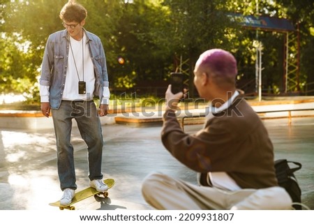 Young stylish boy with phone taking picture of his friend on skateboard , while sitting in lotus pose in skatepark