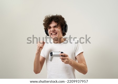Excited caucasian teenage boy celebrate his win in video game with joystick. Curly guy of zoomer generation wear t-shirt and headphones. Youngster lifestyle. White background. Studio shoot. Copy space