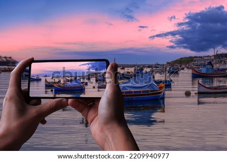 Tourist taking photo of traditional Maltese boats in Marsaxlokk harbor during sunset, Malta