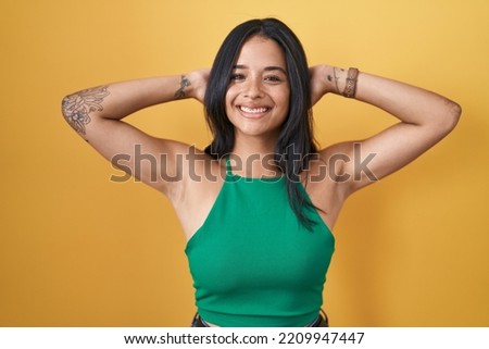 Brunette woman standing over yellow background relaxing and stretching, arms and hands behind head and neck smiling happy 