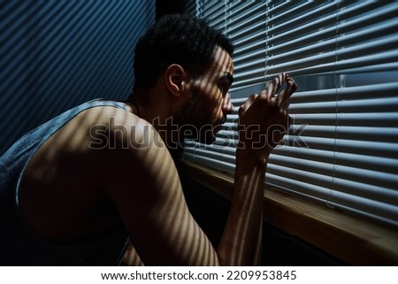 Side view view of young sleepless man looking through venetian blinds on window during sneak peek after someone outdoors Royalty-Free Stock Photo #2209953845
