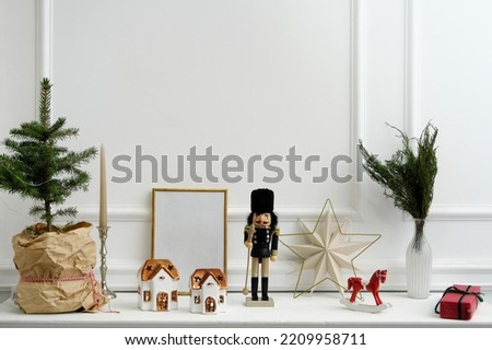 Christmas decor. Nutcracker, candles in the form of houses, a rocking horse and a natural Christmas tree stand on a white shelf. An empty frame for text stands on a shelf. Horizontal photo