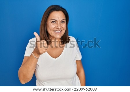 Hispanic mature woman standing over blue background doing happy thumbs up gesture with hand. approving expression looking at the camera showing success. 