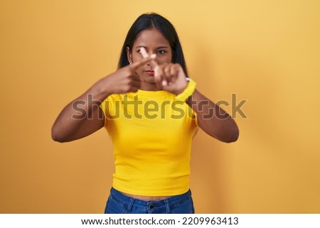 Young indian woman standing over yellow background rejection expression crossing fingers doing negative sign 