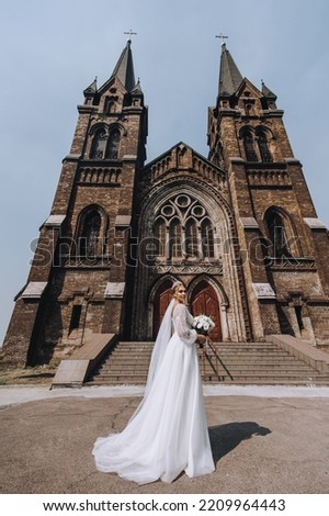 A beautiful, smiling bride in a white long dress with a diadem on her head, with a bouquet, stands against the background of an old brick temple, church. Wedding photography, portrait.