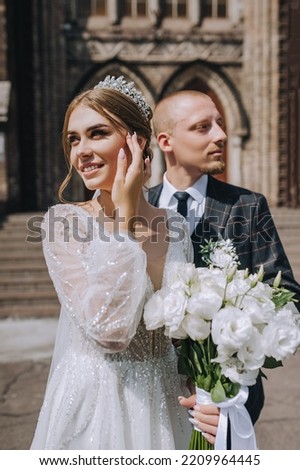 A stylish groom and a beautiful bride with a bouquet stand outdoors in sunlight against the backdrop of a brick temple, building. wedding photography. Royalty-Free Stock Photo #2209964445